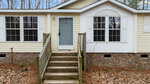 A beige, single-story house with white trim and a gray front door, located at 1188 Sims Bridge Road, Kittrell. The entrance features a short set of brick steps and a railing. Tall windows flank the door, and the house is surrounded by a wooded area with fallen leaves on the ground.
