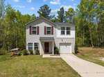 A charming two-story house at 4009 Shady Brook Drive features light gray siding, red shutters, and an attached single-car garage. The grassy front yard has a concrete driveway and sidewalk leading to the front door, with a bench and umbrella to the left. Surrounded by trees, this Kittrell home is a serene haven.