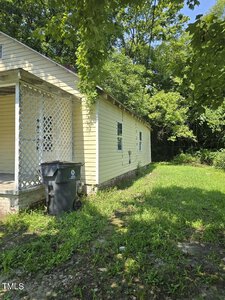 A small, light yellow house sits amid lush green foliage. The side porch has white lattice, and a black garbage bin is placed nearby. The lawn is overgrown, and trees provide shade, suggesting a rural or suburban setting.