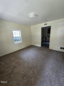 A small, empty room with beige carpet and light-colored walls. A white-framed window allows natural light in from the left wall. A narrow doorway leads to a kitchen area with visible cabinets and a water heater. A ceiling light fixture is centered on the ceiling.
