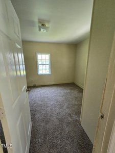 A small, empty room with beige walls and a gray carpeted floor. A window with a white frame is centered on the far wall, letting in natural light. The door is open, allowing a view into the space. A ceiling light fixture is visible.