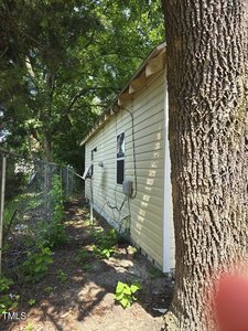 A side view of a yellow house with a tree in the foreground. The house has a beige exterior with visible piping and electrical cables attached. There is a fence along the side of the house, and surrounding greenery, including trees and plants.
