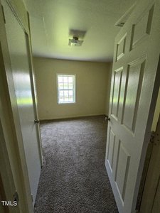 A small, empty room with beige walls and a carpeted floor. A single window lets in natural light. The room is viewed through an open door, and a light fixture is attached to the ceiling.