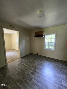 A small, empty room with light gray wood flooring and beige walls. The room has a window and a door opening into another room with a carpeted floor. There is a wooden cabinet mounted on the wall above the window. Sunlight streams in through the window.