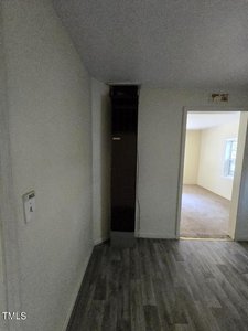 A corner of a room with light-colored walls and wood-patterned vinyl flooring features a tall, narrow heater along the wall. An open doorway leads into another brightly lit room with carpeted flooring and a window. A thermostat is mounted on the wall.