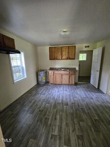 A small kitchen with wooden cabinets and a counter, a sink, and a hot water heater. The room has grey vinyl flooring, a window on the left wall, and a door on the right leading to another room. The walls are painted white, and there is a ceiling light fixture.