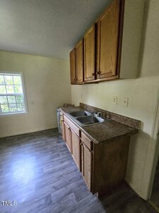 A small kitchen with a sink and counter, wooden cabinets above and below, and a single window on the left wall letting in natural light. The room has light-colored walls and a gray wood-style floor.