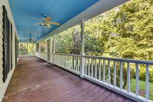 A spacious porch with a blue ceiling and wooden flooring, bordered by a white railing. Two ceiling fans with lights hang from above. The porch offers a view of lush green trees and a grassy yard on the right side.