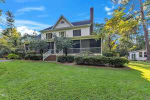 A two-story house with a gable roof, large screened-in porch, and brick chimney set against a backdrop of tall trees at 2055 Fernwood Way in Henderson. The yard is well-maintained with lush green grass and shrubbery, and a small outbuilding is visible in the background to the right.