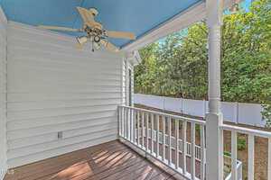 A small outdoor balcony with white railing and trim, featuring a ceiling fan attached to the blue ceiling. The space overlooks a fenced backyard with trees. The balcony has wooden flooring and is attached to a white house with horizontal siding.