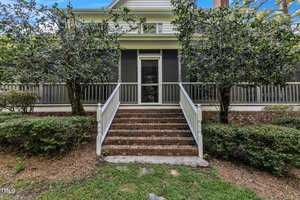 A house with a screened-in porch featuring a central staircase with brick steps leading up to it. The porch has white railings, and the house exterior is light-colored. Lush greenery and bushes surround the area, enhancing the natural aesthetic.