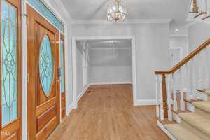 A spacious foyer with a polished wooden floor leads to an adjacent empty room. The area features a wooden front door with frosted glass panels, an elegant chandelier on the ceiling, and a wooden staircase with white railings on the right side.