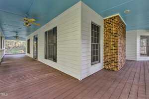 A spacious wraparound porch with a wooden floor and blue ceiling extends around a white house with brick accents. The porch features multiple windows and ceiling fans, creating a welcoming outdoor space. Trees are visible in the background.