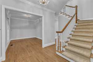 A spacious interior featuring a carpeted staircase with a wooden handrail on the right and a hardwood-floored room with large windows on the left. The walls are painted light gray, and both rooms are illuminated by elegant ceiling light fixtures.