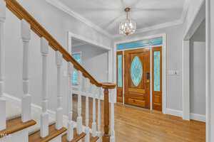 A brightly lit foyer with a wooden staircase on the left, leading up to the second floor. The front door features stained glass windows with intricate designs, and a chandelier hangs from the ceiling. White walls and a hardwood floor complete the elegant look.