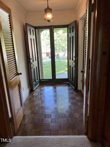A hallway with wooden parquet flooring and a carpeted area leading to double front doors with glass panels, opening to a view of a green lawn. The interior features natural wood accents, including louvered closet doors and a hanging light fixture in the ceiling.
