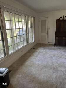 A living room with large windows providing ample natural light. The space is carpeted, and there is a closed white door with a louvered section. A dark brown wooden cabinet stands against the far wall. A black item is placed near the window.