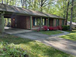 A single-story brick house at 307 Yowland Road in Henderson features a green front porch, surrounded by trees and greenery. The front yard boasts a manicured lawn and flowering bushes by the entrance. A paved driveway leads to the house, which has windows adorned with green shutters.