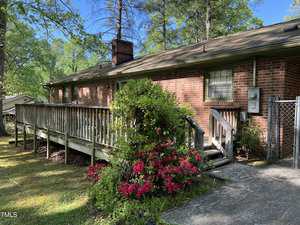 A single-story brick house with a sloping wooden ramp leading to the front entrance. The yard features well-maintained greenery, including a prominent bush with pink flowers near the walkway. Trees surround the property, and a chain-link fence is partially visible.