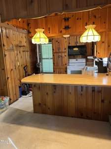 A kitchen with wood-paneled walls and cabinets, a wooden countertop with a bar, two hanging pendant lights, a stove, a microwave, and various small appliances on the counter. A plastic bin filled with items is visible to the left.