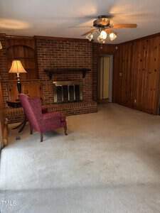 A cozy living room with a brick fireplace, wooden paneling on the walls, and beige carpet. A maroon armchair is placed next to a wooden side table with a lamp. A ceiling fan with lights hangs from the white ceiling. Shelving is built into the wall near the fireplace.