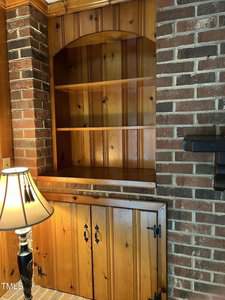 A wooden built-in shelving unit is set within a brick wall. The lower section features closed cabinet doors, while the upper section has open shelves. A lamp with a beige shade stands next to the unit. The setting appears to be a cozy indoor space.