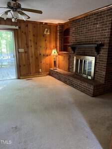 A living room with wood-paneled walls, a ceiling fan with light, and a large brick fireplace with a mantle. There is a small table lamp on the built-in shelves next to the fireplace. A sliding glass door leads to a deck outside. The carpet is light-colored.
