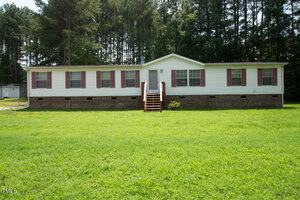 A one-story white house with brown shutters and a brick foundation stands in front of a grassy lawn at 20 Shearin Lane. A set of wooden steps leads to the front door, and tall trees are visible in the background creating a wooded area. Welcome to Henderson's charming homestead!