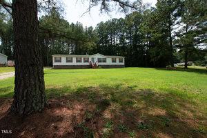 A single-story white house with a brick foundation and brown shutters stands in the middle of a spacious, grassy lawn. A set of wooden stairs leads to the front door. Tall trees line the background, providing a natural, wooded setting.
