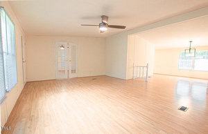 A spacious living room with light wood flooring, neutral-colored walls, and a ceiling fan with a light fixture. Large windows allowing natural light fill the room. French doors lead to an adjacent room, visible through a wide open doorway.