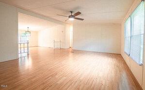 Spacious, unfurnished living room with light hardwood flooring, white walls, and large windows with blinds. A ceiling fan hangs in the center. An adjoining room is visible with similar decor and a decorative railing dividing the spaces. Natural light fills the rooms.