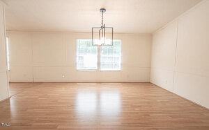 Empty, well-lit room with light-colored wood flooring, white walls, and a modern black metal chandelier with exposed bulbs hanging from the ceiling. Two large windows with blinds are centered on the far wall, allowing natural light to flood the room.