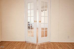 A set of white French doors with glass panels, partially open, leads into a room with wooden flooring and minimal decor. A chair and ceiling fan are visible in the adjacent room through the glass panes.