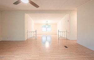 A spacious, empty room with light wooden flooring and white walls. There is a ceiling fan in the foreground, a recessed area with white railing, and a window with sheer curtains allowing natural light in the background.