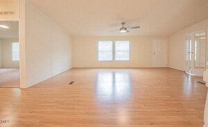 A spacious, empty living room with light-colored hardwood flooring, a ceiling fan, and three windows providing natural light. There's a doorway on the right that leads outside, and a set of French doors on the right that lead to another room.