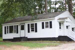 A small white house with black shutters and a front porch is surrounded by trees. The house has a dark foundation and white steps leading up to the front door. The surrounding grassy area shows a dirt path leading to the house.