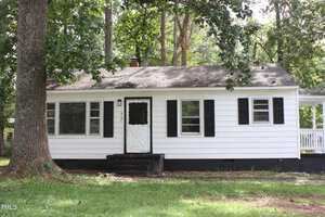 A small, single-story white house with black shutters and a black front door is surrounded by tall trees and greenery. The house has a modest front porch and windows on either side of the door. The lawn in front is grassy and shaded by trees.