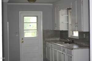 A small kitchen with gray walls and cabinets. It features a sink under a window, a countertop, and a door with a window panel leading outside. The kitchen has a gray tiled backsplash and white trim.
