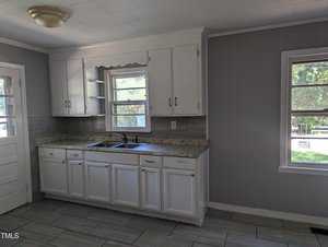 A kitchen with white cabinetry, a double sink under a window, granite countertops, and gray tile flooring. Natural light streams in through two windows, highlighting the clean, minimalist space. A door is visible on the left side of the room.