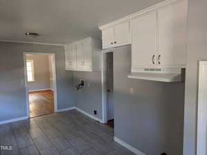A room with light gray walls, white ceiling, and tiled gray flooring. White cabinets are mounted on the upper walls, and a stove hood is affixed above a countertop area. An open doorway leads to an adjoining room with wooden floors and a window, allowing natural light.