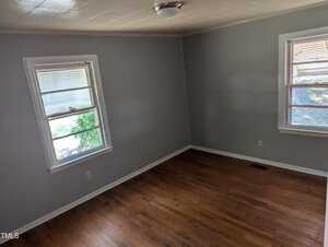 A room with light gray walls, two windows letting in natural light, and a ceiling light fixture. The floor is wooden, and the room appears empty, with no furniture or decorations.