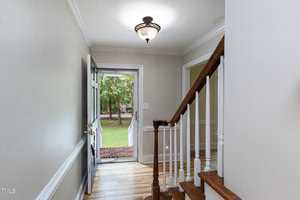 A well-lit entryway of a home featuring a staircase with wooden steps and white railings on the right. The front door, which is open, reveals a view of a green lawn and trees outside. The walls are painted light gray, and there is a light fixture on the ceiling.