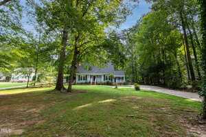 A charming white house with dark shutters and a steep roof sits surrounded by tall, leafy trees. The front yard is spacious, with a lush lawn and a long driveway to the right, leading to a side entrance. Sunlight filters through the trees, casting serene shadows.