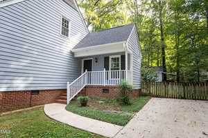 A small, quaint house with light blue siding, red brick foundation, and a white front porch. The house has a black front door, white railing, and is surrounded by trees. A curved concrete pathway leads from the driveway to the porch.