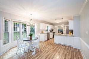 A bright kitchen and dining area with wooden flooring. The dining area features a round table with four white chairs by a large bay window. The kitchen has white cabinets, stainless steel appliances, and a central island with a fruit bowl and other decorations.