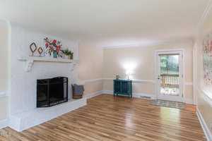 A well-lit living room with a white brick fireplace on the left, adorned with a floral arrangement and decorative items on the mantel. The room has hardwood floors, a blue console table with a lamp near the glass door leading to a wooden deck outside.