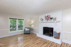 A bright living room with light-colored walls and wooden flooring. The room features a white brick fireplace with a decorated mantel, a patterned armchair next to a floor lamp and side table, and a large window allowing natural light to fill the space.
