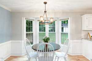 A cozy dining nook featuring a round wooden table with a small potted plant centerpiece and four white chairs. Natural light streams in from a bay window with views of a lush, green backyard. The walls are painted light gray, complemented by white trim and cabinets.