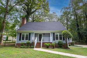 A charming white house with a dark roof, brick foundation, and blue shutters, nestled among tall trees. A curved path leads to the front steps, with a grassy lawn and a neatly trimmed hedge lining the entrance.