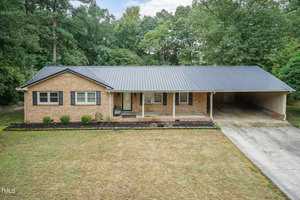 A single-story brick house with a black metal roof features a covered front porch and an attached carport. It has two large front windows and a neatly maintained front yard with bushes along the house and a concrete driveway. Trees are seen in the background.